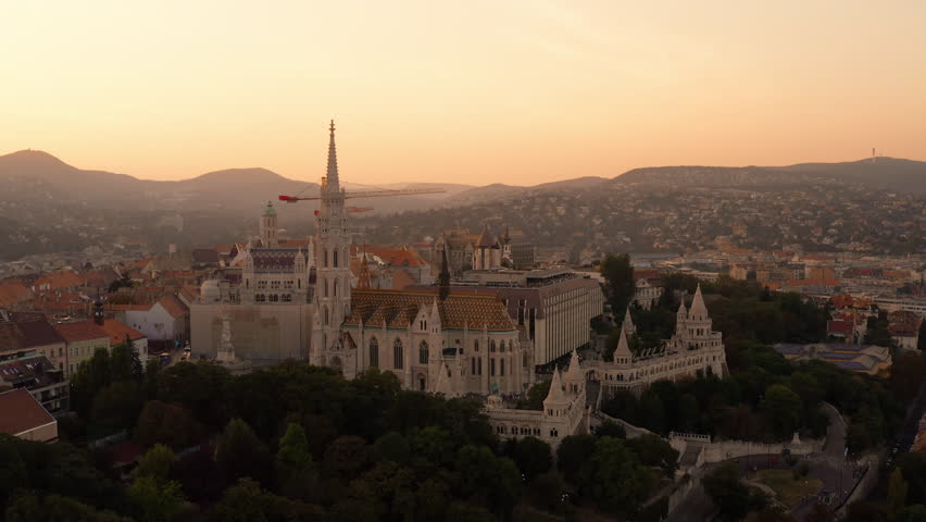 Panoramic view of Buda Castle Hill during sunset. Burning orange sunset skies over the Buda Hills. Cranes indicating the ongoing renovation of Buda Castle. Matthias Church, Fisherman's Bastion Hungary Royalty-Free Stock Footage #3471955387