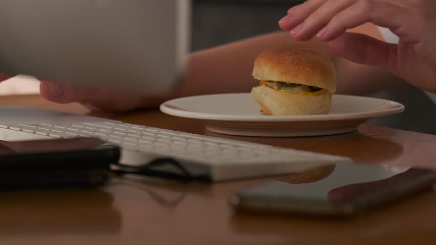 Young woman working at a computer in the office snacks on a sandwich without looking up from her work, chews bread with appetite, sitting at the table opposite the monitor, close up.