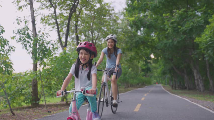 Asian young mother and daughter riding bicycle outdoors in the garden. Happy family, attractive mommy in sportswear feeling happy and relax, exercise in public park with adorable kid girl for health.
