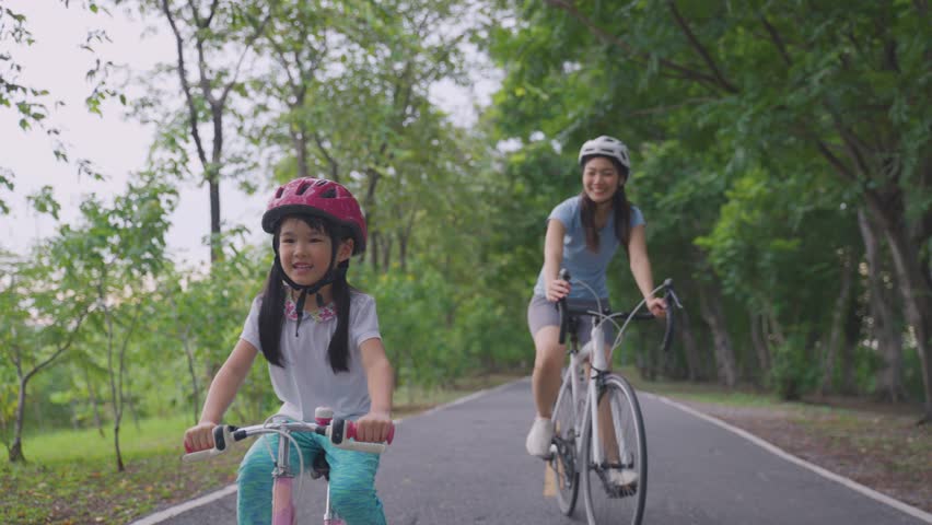 Asian young mother and daughter riding bicycle outdoors in the garden. Happy family, attractive mommy in sportswear feeling happy and relax, exercise in public park with adorable kid girl for health.