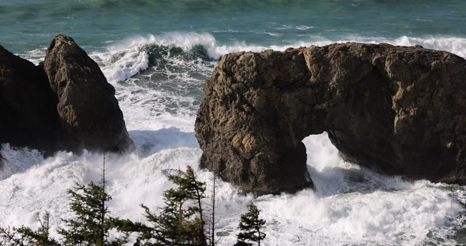 A rocky shoreline with a large wave crashing into a rock formation. The waves are high and powerful, creating a sense of energy and movement. The rocks are scattered throughout the scene