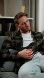 Vertical video Close-up shot of a happy blond man with stubble in a checkered shirt playing a black musical instrument the ukulele while sitting on a gray sofa in a modern apartment during the day