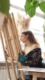 Female artist drawing in light art studio.Beautiful young woman sitting in front of wooden easel on vertical video. Blonde girl in black floral dress in creative process