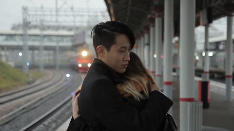 Young man is hugging woman standing at railway station outdoors. multiethnic Enamored people cuddle warmly, embracing each other with hands and touching with bodies. Korean guy dressed in stylish