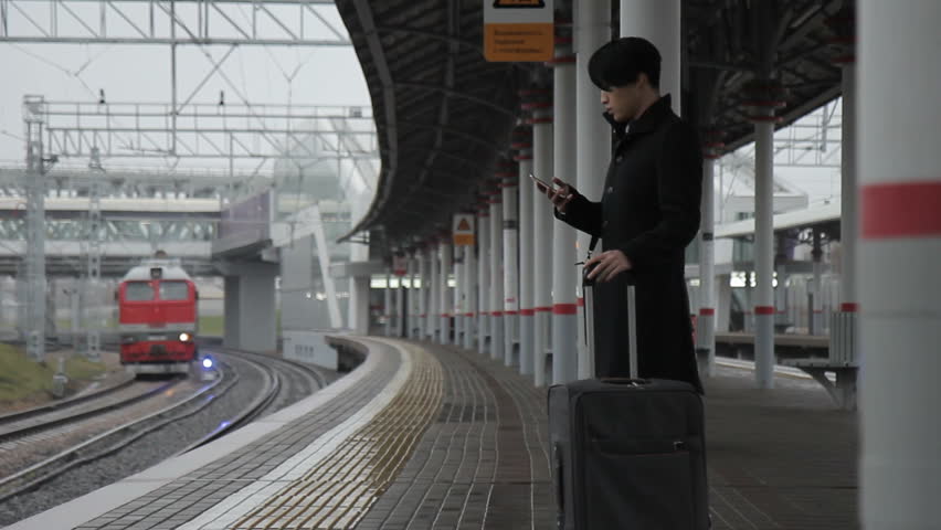 Asian man with suitcase and phone is standing on railway platform. Man is waiting for train to travel and typing text on a smartphone with luggage.