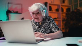 Humorous 80-year-old woman in headset playing multiplayer game on laptop at home