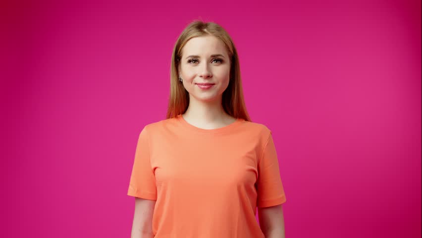 Smiling Young Woman in Orange Shirt Waving Hello Against Pink Background