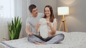 Beautiful pregnant woman and her handsome husband holding bodysuit for future baby and rejoicing man gently kissing his loving woman enjoying happy pregnancy and child expectation - Powered by Shutterstock - Get 15% off with code: PIKWIZARD15