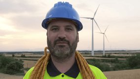  Headshot of male maintenance worker standing in wind turbine farm field with climbing equipment: helmet and rope. 4k video Looking at camera. Engineering and renewable energies.
