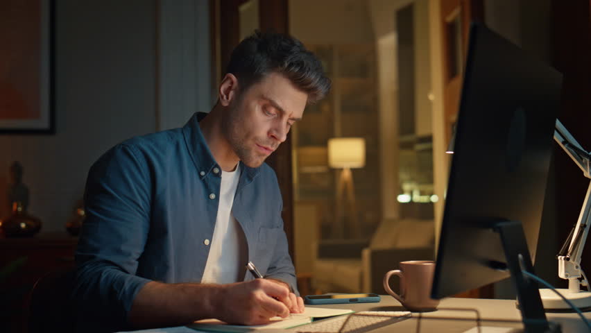 Online man studying pc night home looking screen closeup. Serious student making notes in copybook sitting desk with computer. Focused freelancer taking internet course watching display video lesson