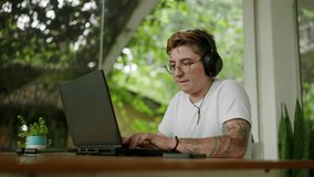 Transgender editor with headphones works on video project in coworking space. Inked professional edits film on laptop, comfortable open office environment. Inclusive workplace, adaptive tech use.