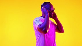Portrait of smiling positive African-American man listening to music in headphones against yellow studio background in neon light. Concept of human emotions, casual fashion, lifestyle