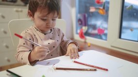baby girl draws while sitting at table by the window at home. happy family kid concept. baby daughter learns to draw with pencils on a sheet of dream paper indoors. development of fine motor skills - Powered by Shutterstock - Get 15% off with code: PIKWIZARD15