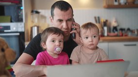 father working from home remotely with two baby in his arms. pandemic remote work business concept. fun father tries to work at home in kitchen, baby children interfere sitting on their hands