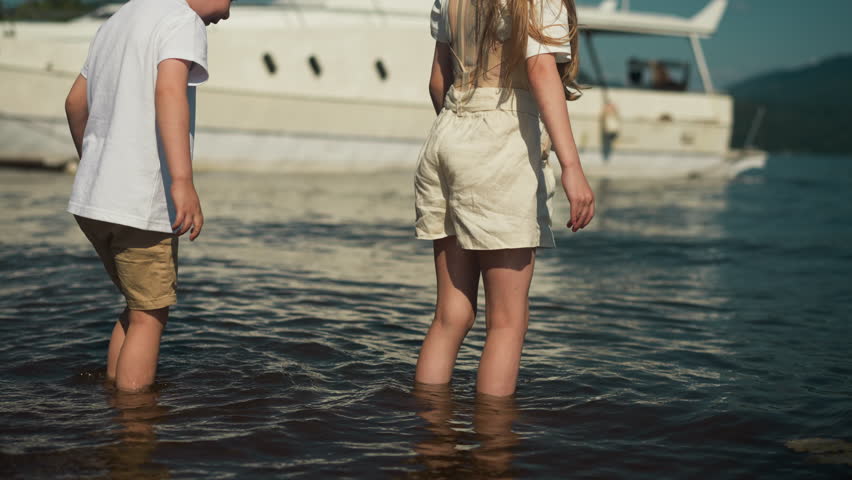 Curious girl and boy walk knee-deep into sea against modern yacht. Recreation in beauty of nature. Spending time together and enjoying environment in summer