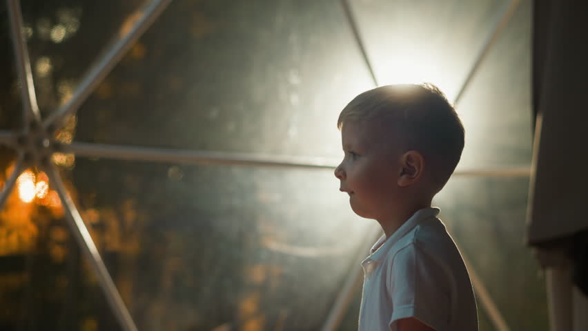 Small kid stands against light of lantern behind tent window. Tired cute preschooler goes to rest in evening at resort on glamping area slow motion backside view closeup