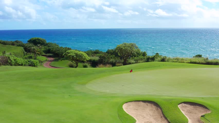 Dream green golf course at ocean in Hawaii. Red flag waving on putting green hole. Aerial above green golf course clear transparent blue ocean water. round  shaped sand trap bunker