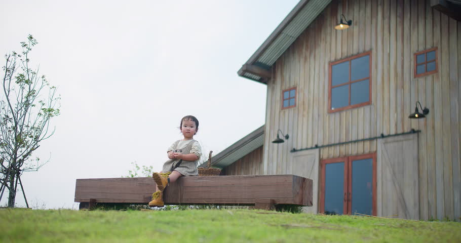 A young child sits on a bench in front of a large wooden house. The child is wearing a dress and yellow boots holding a basket and he is enjoying the outdoors. The scene is peaceful and calm