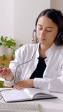 Vertical HD video. Young female doctor taking notes or writing a medical prescription for her patient while having an online conversation on computer video call at hospital