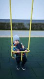 Beautiful Caucasian baby boy wearing warm jacket and cap sits in the swing. Lovely toddler having fun on the playground outdoors. Vertical video.