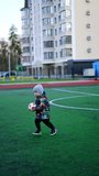 Energetic toddler boy runs carrying a ball in hands. Two-year-old child playing active games outdoors at stadium. Vertical video.