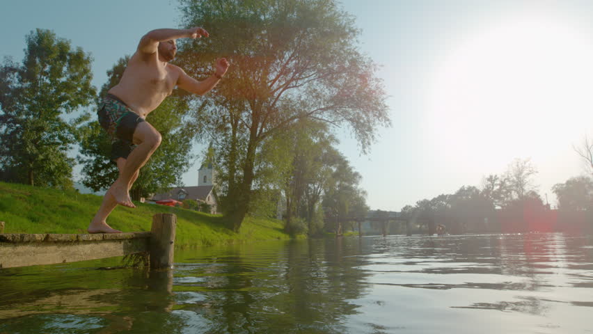 SLOW MOTION, LENS FLARE: Youthful man jumps into water and creates a huge splash. Water droplets fly around when a carefree young guy jumps and plunges into a refreshing river on a hot summer day.