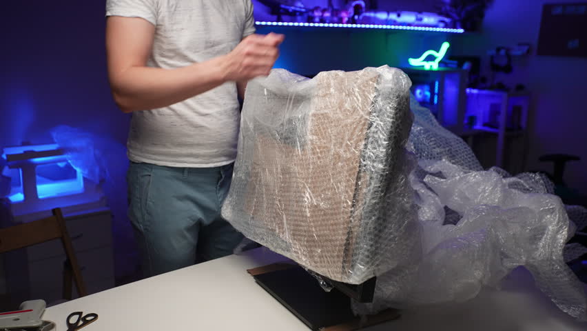 Young man packing desktop monitor or TV in cardboard box and bubble wrap standing at table in dark room during preparation to relocation new apartment. Concept of new life in new home relocation. Royalty-Free Stock Footage #3472778771