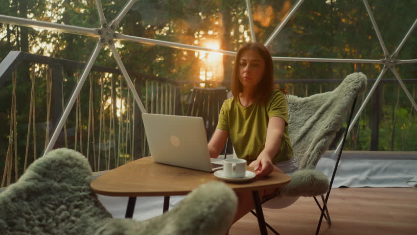 Busy woman drinks tea in cup working in online sphere in tourist house surrounded by forest. Business trip to another country with incredible views at sunset slow motion