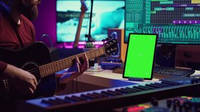 Male artist learning to play guitar on online lesson tutorial, practicing acoustic instrument and looking at phone with greenscreen display. Music producer doing rehearsal guitar play. Camera A.