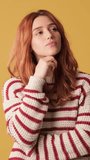 Vertical video, Pensive red-haired woman dressed in sweater thinks something, makes choice on yellow background