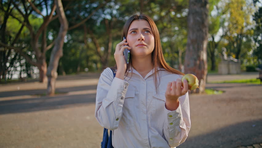 Positive lady talking phone while savoring apple in park. Closeup casual woman have smartphone conversation with nature backdrop enjoying sunny day. Outdoor setting blending technology and leisure
