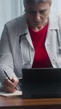 Vertical video. Mature female doctor in white coat work at workplace sit on sofa, writes notes, prepare report, filling patient card, prescribe medications