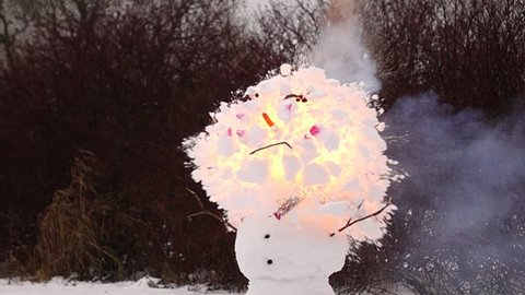 Super slow motion shot of petard explosion inside Snowman head. Fire burst inside top ball and snow fly apart around, destroying one snowball completely. Funny smiling face with pink cheeks blow up