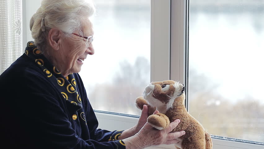 A cheerful positive old woman playing with a soft toy by the window. lonely crazy grandmother