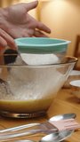 Vertical video. A woman sifts flour into a glass bowl in slow motion, part of the process of making a cake.