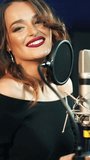 Beautiful playful singer at the professional microphone in audio studio. Attractive woman singing happily into the mic indoors. Female voice. Vertical video