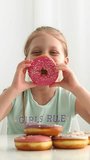 Portrait of a little smiling girl with a delicious donut in her hand, pink chocolate glaze on donuts. Diet and unhealthy food concept. June 7 National Donut Doughnut Day. Vertical video.