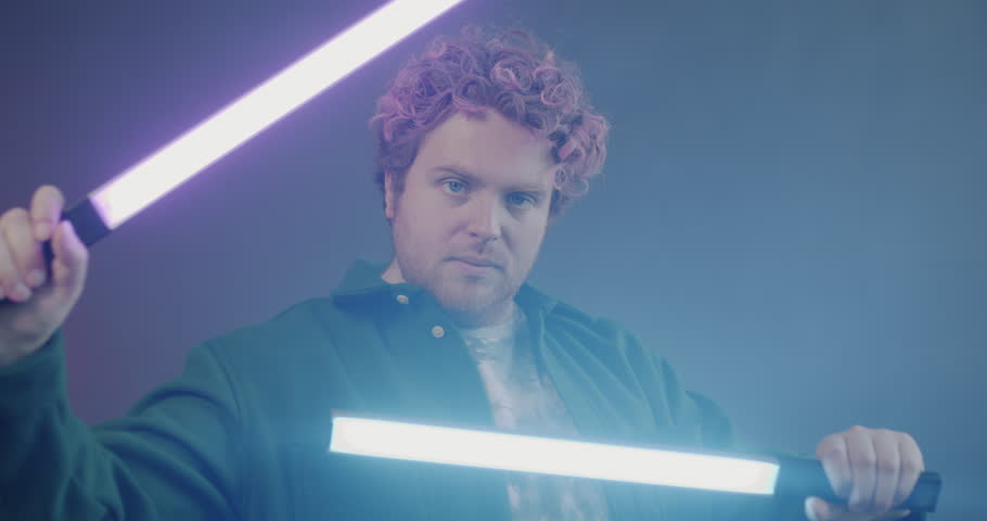 Slow motion portrait of creative young man moving hands with neon light sticks against smoke purple background. Illumination and cyberpunk concept.