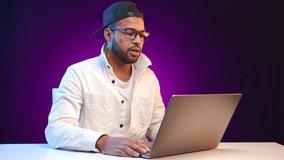 A man in casual attire is fully engaged in a video call, his laptop screen illuminating his focused expression against a vivid purple backdrop. Captured with precision detail. Camera 8K RAW. 