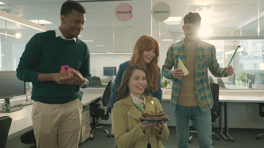 An interracial businesswoman is having a birthday surprise at the office with colleagues. A happy colleague with disability is about to hug her while a birthday girl is holding a cake. Bday at office.