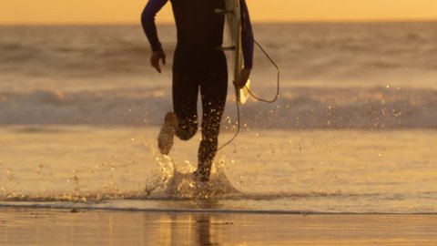 SLOW MOTION: Young surfer running into water to surf breaking waves at golden sunrise. Carefree male surfboarder jogging into ocean to ride waves on his cool surfboard in the beautiful summer sunset.