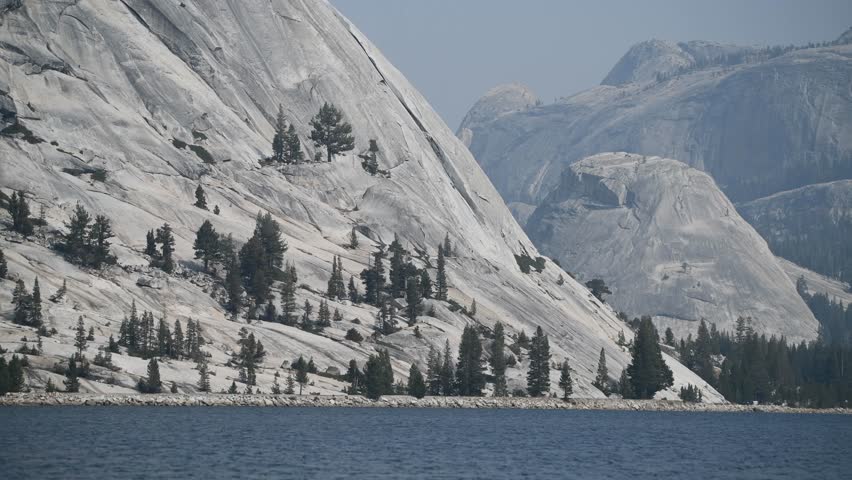 Closeup of the coast of Lake Tenaya and the mountains of gray rocks and patches of trees that surround it. View during the 2021 California wildfires Royalty-Free Stock Footage #3473250043