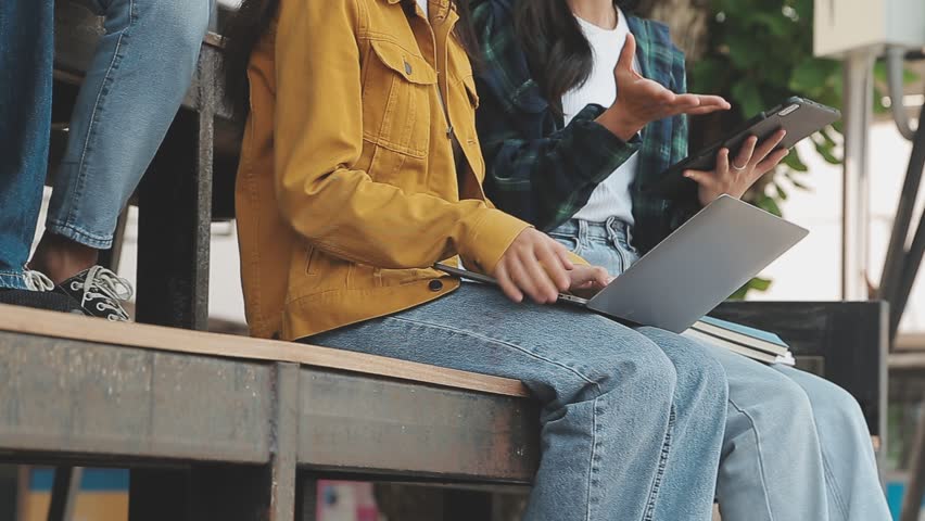 Group of happy young Asian college students sitting on a bench, looking at a laptop screen, discussing and brainstorming on their school project together.