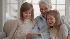 Grandmother with granddaughters resting. Attractive relaxed older adult granny embracing watches cartoons on smartphone with children two twins leisure rest at home. Family, generation concept.