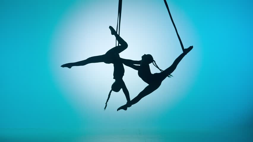 Silhouette of two female acrobats isolated on blue neon background. Girls aerial dancers performing acrobatic flying on ropes.