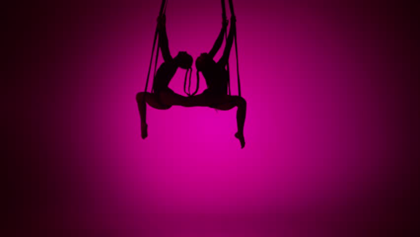 Silhouette of two female acrobats isolated on pink neon background. Girls aerial dancers performing mirrored element on ropes.