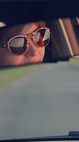 smiling woman driving, fashionable sunglasses reflection, looking through mirror, joyous driving experience, pleasant road trip, casual driving attire, enjoying sunny day, relaxed seating position Royalty-Free Stock Footage #3473455395
