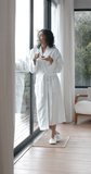 Vertical video of biracial woman wearing bathrobe drinking tea at home, slow motion. Lifestyle, relaxation, self care and domestic life, unaltered.