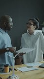 African American renewable energy engineer holding business papers and explaining points of project to young female coworker during day in the office. Vertical clip