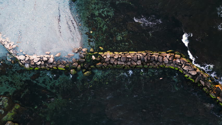 Seagulls soar along breakwater built out from grey white beach, drone top down